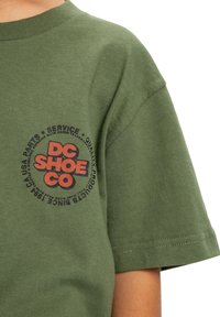 Olivově zelené tričko s krátkým rukávem vyrobené z bavlny, s kulatým logem v červené a černé barvě na levé straně hrudi se textem kolem něj.