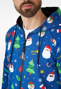 Sudadera con cremallera azul que presenta patrones festivos de Santa, árboles de Navidad, copos de nieve y bastones de caramelo, fabricada en una suave tela texturizada.