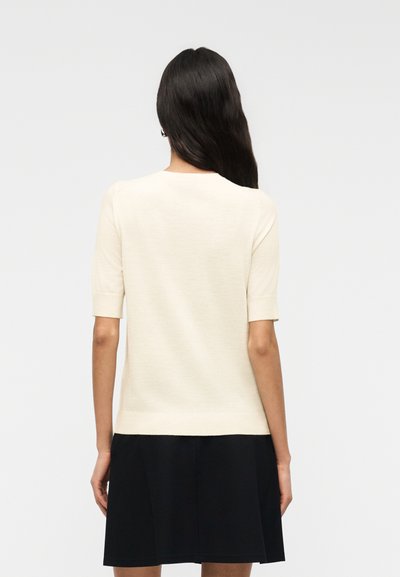 Femme aux longs cheveux foncés portant un haut crème uni à manches courtes et une jupe noire, vue de dos sur un fond clair uni.