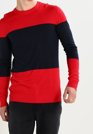 Homme portant un pull à manches longues avec des blocs de couleurs horizontaux rouge et bleu marine, associé à un pantalon foncé, debout devant un fond blanc uni.