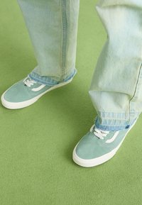 Miętowe zamszowe sneakersy z białymi akcentami, w połączeniu z jasnoniebieskimi, lekko postrzępionymi dżinsami, na zielonym tle.