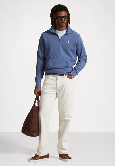Sudadera azul con cremallera, camisa azul claro debajo, pantalones blancos, bolsa marrón y zapatos marrones. Unas gafas de sol notables completan el look.