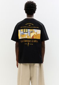 Fekete, pamut anyagú póló, amelynek hátulján van Gogh "La Chambre à Arles" című művének sárga és kék nyomtatott mintája található.