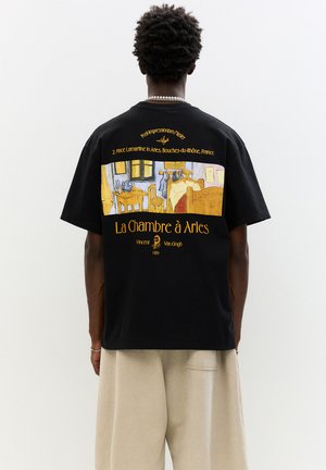 Sort T-shirt lavet af bomuld med et trykt design af Van Goghs "La Chambre à Arles" i gul og blå på bagsiden.