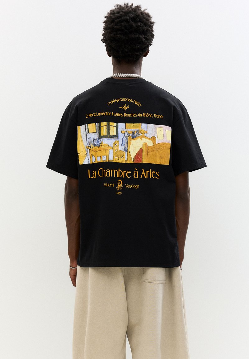 Fekete, pamut anyagú póló, amelynek hátulján van Gogh "La Chambre à Arles" című művének sárga és kék nyomtatott mintája található.