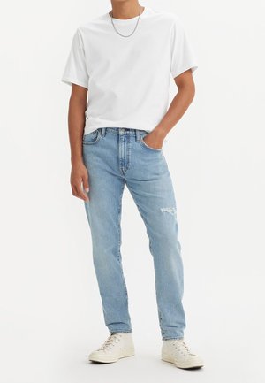 Jean en denim bleu clair avec un genou déchiré, associé à un t-shirt blanc uni et des baskets blanches, présentant un col rond et des manches courtes.