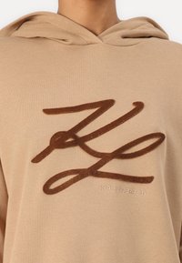 Hanorac bej cu un mare logo brodat maro, având un design minimalist și glugă cu șnur. Textul spune "KARL LAGERFELD."