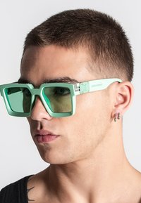 Gafas de sol grandes de color verde menta con lentes cuadrados, montura de plástico elegante y branding sutil en la patilla. Acabado reflectante ligero visible.
