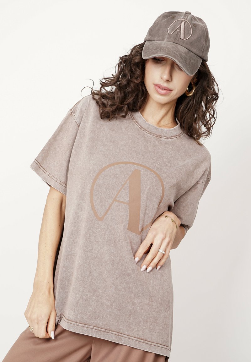 Brązowy oversize t-shirt z krótkimi rękawami i dużym beżowym logo "A" na przodzie. W zestawie z dopasowaną brązową czapką z tym samym logo.