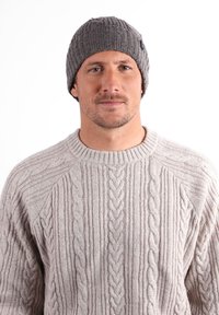 Bonnet en tricot gris avec une texture côtelée. Le modèle porte un pull en maille torsadée beige clair avec un col rond. Design simple et polyvalent.