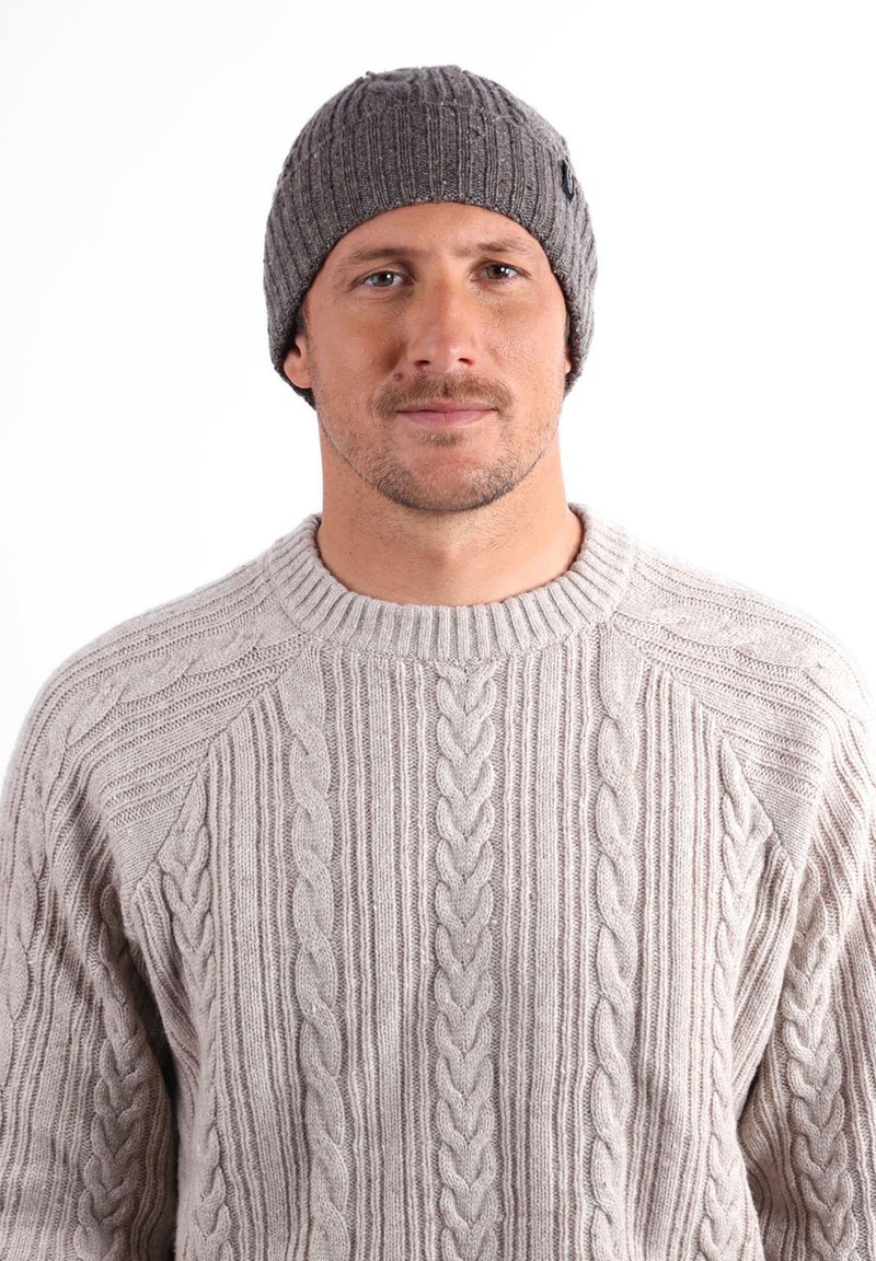 Bonnet en tricot gris avec une texture côtelée. Le modèle porte un pull en maille torsadée beige clair avec un col rond. Design simple et polyvalent.