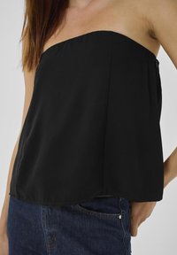 Schwarzes trägerloses Top aus glattem Stoff, mit geradem Ausschnitt und fließendem Design. Kombiniert mit blauen Jeans.
