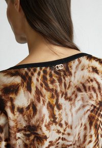Durchscheinendes Oberteil mit Animal-Print und schwarzem Besatz, das am Ausschnitt ein dezentes goldenes Logo-Detail sowie einen leichten, strukturierten Stoff aufweist.
