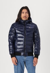 Mornarski puffer jakna sa sjajnom završnom obradom, sa patentnim zatvaračem sprijeda, kapuljačom i rebrastim manžetama. Sadrži kviltani dizajn i mali logotip na prsima.