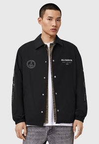 Giacca nera con chiusura frontale a scatto, tasche laterali e ricamo di un simbolo della pace. Presenta testi bianchi a contrasto e dettagli grafici sulle maniche.