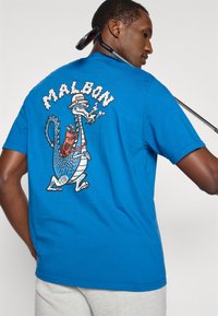 Mėlyna medvilninė marškinė su drakono grafika, dėvinčio kepurę ir rūkymo detalėmis ant nugaros. Trumpomis rankovėmis, laisvas fasonas.