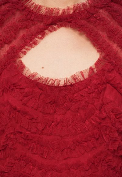 Raudona, raukšlėta palaidinė iš permatomo audinio, turinti skylutę apykaklėje ir tekstūruotą, sluoksniuotą dizainą.