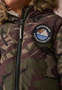 Zaļā un brūnā toņu kamuflāžas jaka ar mākslīgās kažokādas kapuci, ar rāvējslēdzēja aizdari un apaļu izšūtu uzpatch ar kalnu dizainu.