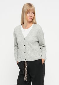 Grijze cardigan met een V-hals, knoopsluiting aan de voorkant, geribbelde zakken en omgeslagen mouwen. Combinatie met zwarte broek met een luipaardprint trekkoord.