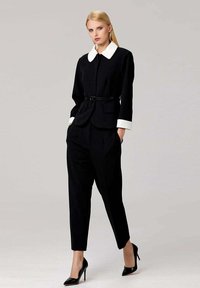 Costume noir avec un col blanc, veste sur mesure, taille ceinturée et pantalons fuselés, associés à des escarpins noirs. Texture du tissu lisse.