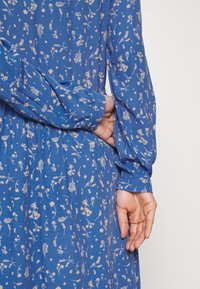 Blå mönstrad klänning med långa ärmar som har en delikat blommönster i krämfärg. Tyget ser mjukt och strukturerat ut. Handen placerad på midjan.