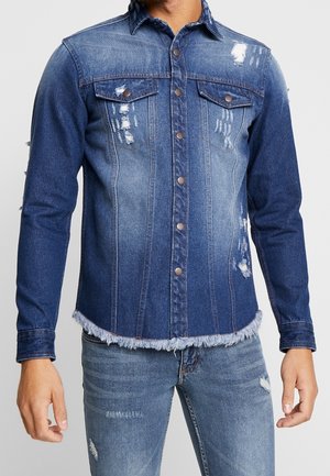 Muž nosící modrou džínovou bundu s knoflíky vpředu a třepeným lemem, doplněnou světle modrými roztrhanými džínami, viditelný trup.