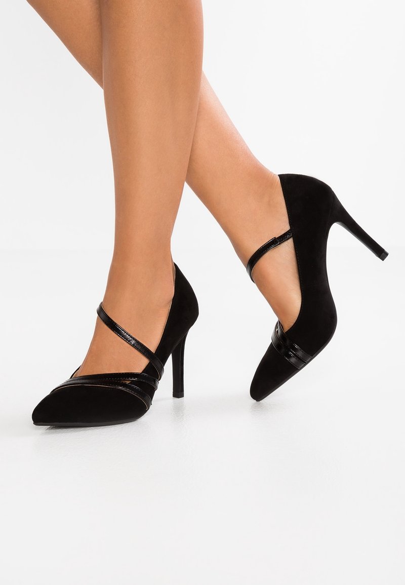 Escarpins en daim noir à bout pointu, avec des brides noires brillantes traversant l'avant et la cheville, posés sur une surface blanche lisse.