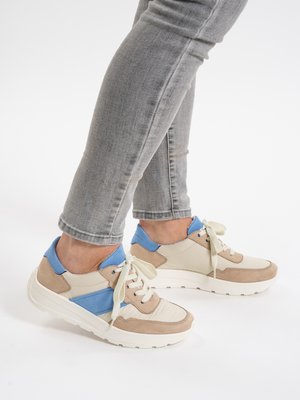 Person wearing beige, tan, and blue sneakers with white laces, paired with gray skinny jeans against a plain white background.