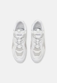 Zapatillas blancas con detalles de malla y cuero. Presenta un diseño robusto, detalles en gris, cierre de cordones en la parte delantera y el logo de Versace en la lengüeta.