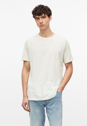 Jeune homme aux cheveux bouclés portant un t-shirt blanc uni à manches courtes et un jean bleu clair, debout avec une main dans la poche.