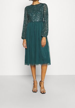 Szmaragdowa sukienka z cekinowymi, długimi rękawami i górą, plisowana spódnica z tiulu, noszona z czarnymi sandałami na wysokim obcasie z paskami na kostkę i palce.