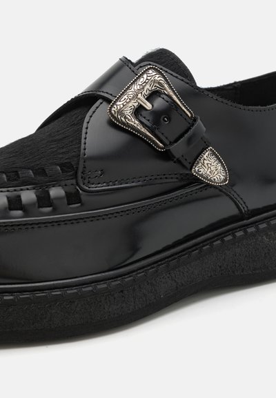 Chaussure en cuir noir avec une finition brillante, dotée de détails tissés, d'une boucle accentuée d'argent et d'une semelle noire texturée.
