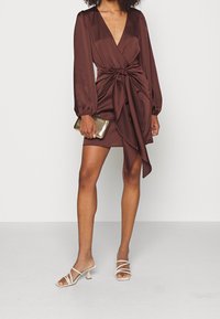 Brun satängminikjol med lång puffärmar, djup V-ringning, knutdetalj på framsidan och asymmetrisk fåll. Kombinerad med en guldfärgad clutch och remsandaler.