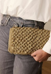 Gouden clutch van gevlochten materiaal met een gewoven textuur, rechthoekige vorm en een ritssluiting, tegen een achtergrond van grijze denim vastgehouden.