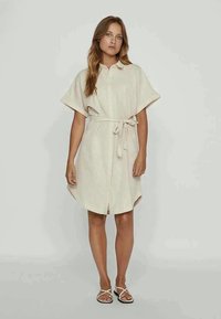 Žena stoji u bež haljini do koljena s kratkim rukavima i pojasom u struku, uparena s bijelim sandalama.