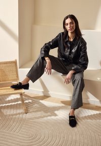 Zwarte leren jasje over grijs gemêleerde broek, gecombineerd met zwarte loafers met gouden accenten. Neutrale achtergrond met een geweven tapijt.