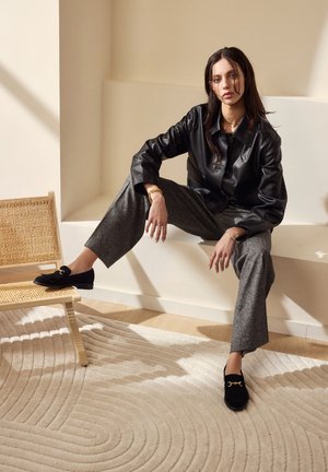 Black leather jacket over gray patterned trousers, paired with black loafers featuring gold hardware. Neutral background with textured rug.