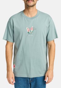 Lys teal bomulds-T-shirt med broderet sommerfugledesign i pink, blå og sort, med blomsterbroderi detalje ved kanten.