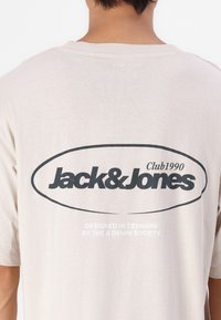 Lys beige t-skjorte med korte ermer, med et stort svart ovalt logo "Jack&Jones Club1990" og hvit tekst nedenfor. Myk innvendig tekstur.