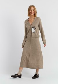 Cardigan in lana beige con chiusura frontale a bottoni e un design di pecora in stile fumetto. Abbinato a una gonna lunga coordinata e mocassini scuri.