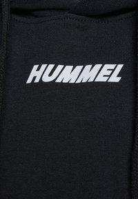 Zwarte stof met witte vetgedrukte hoofdletters "HUMMEL" gedrukt in een schuine, sportieve lettertype.