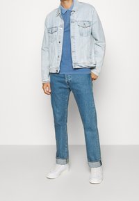 Veste en jean bleu clair portée sur un polo bleu, associée à un jean droit bleu moyen et des baskets blanches. Design simple et coupe décontractée.