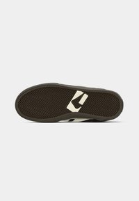 Semelle de chaussure avec motif en chevrons brun foncé et logo rectangulaire blanc près de la voûte plantaire sur fond blanc.