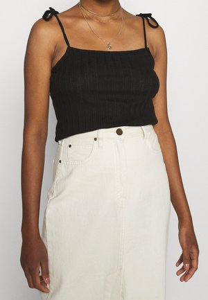 Femme portant un crop top noir sans manches côtelé avec bretelles nouées et une jupe blanche taille haute avec poches avant et fermeture par boutons.