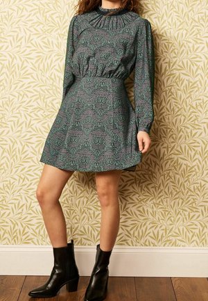 Robe à manches longues, à motif floral vert, avec un col froncé et une taille élastique, associée à des bottines noires à talons.