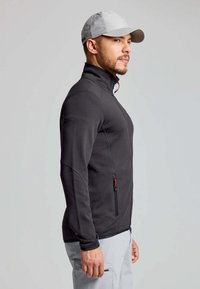 Mann mit einer hellgrauen Mütze, schwarzer Reißverschlussjacke mit roten Reißverschlussanhängern und hellgrauen Hosen, der im Profil vor einem einfarbigen Hintergrund steht.