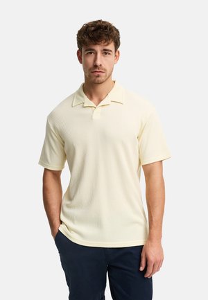 Uomo con capelli castano chiaro che indossa una polo a maniche corte color crema e pantaloni scuri, in piedi con una mano in tasca.