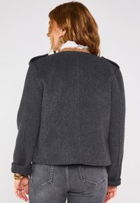 Veste en laine grise au design structuré, avec épaulettes et manches retroussées. Elle présente un tissu texturé et une coupe légèrement raccourcie.