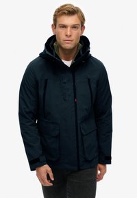 Chaqueta aislante azul marino con capucha, con cremallera frontal, dos grandes bolsillos y un logotipo texturizado en el pecho. Diseño informal.