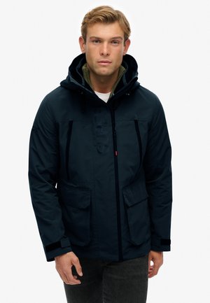Navyblauwe geïsoleerde jas met een capuchon, voorzien van een rits aan de voorkant, twee grote zakken en een gestructureerd logo op de borst. Casual ontwerp.